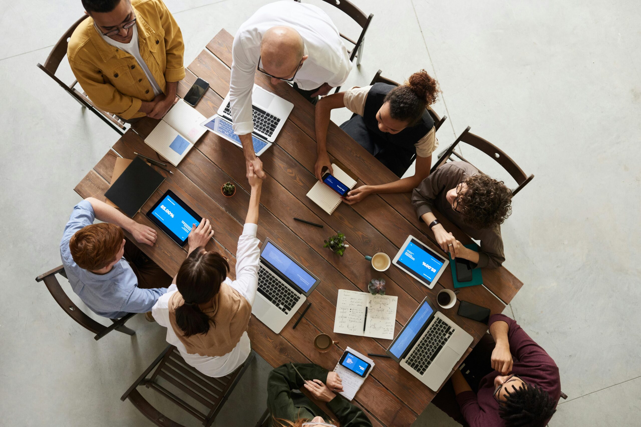 A group of people sitting around a wooden table with laptops and notebooks, collaborating in a meeting as two individuals shake hands.
